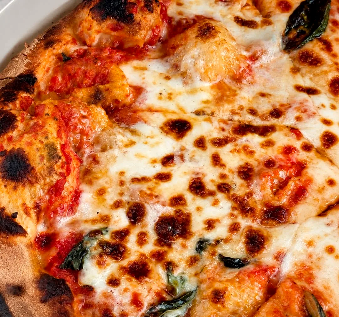 Close crop of a wood-fired margherita: blistered crust, milky mozzarella, basil