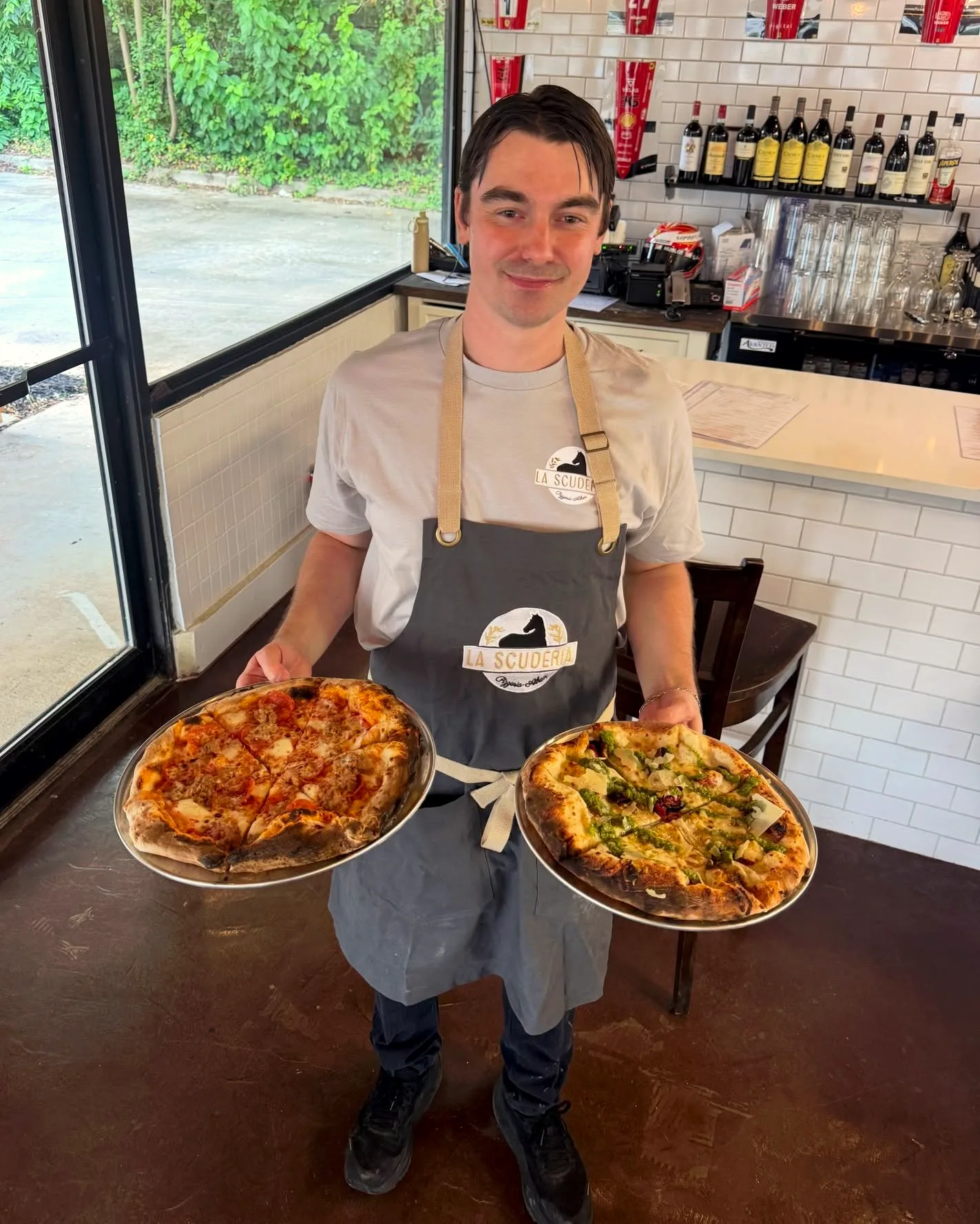 A member of the La Scuderia team carrying two finished pizzas on steel trays past the bar